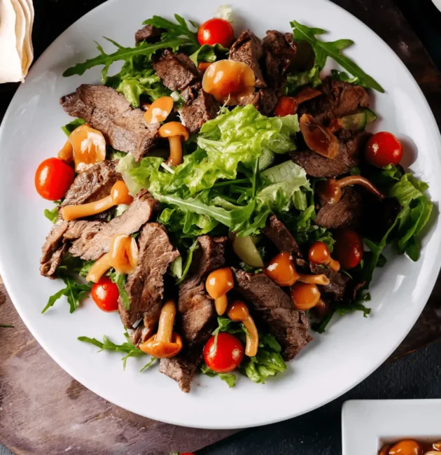 A vibrant lunch plate featuring sliced grilled beef atop a fresh salad with cherry tomatoes, mixed greens, and marinated mushrooms, garnished with a light dressing.