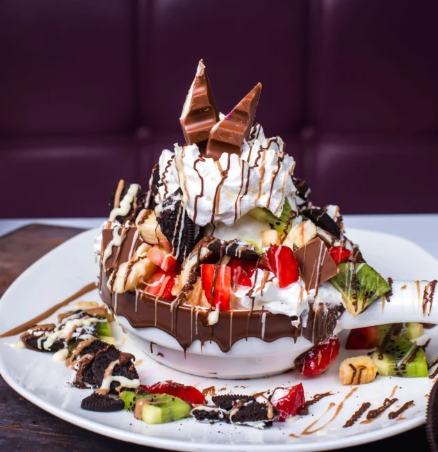 A decadent dessert featuring a chocolate-dipped waffle bowl overflowing with fresh fruit, whipped cream, Oreo crumbles, and chocolate drizzles, topped with chocolate bars.