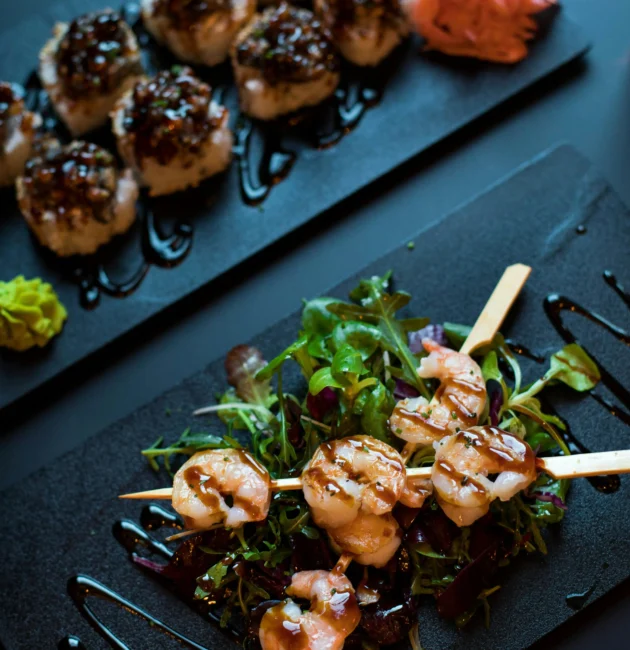 A stylish presentation of appetizers featuring grilled shrimp skewers on a bed of mixed greens and bite-sized rice balls topped with savory glaze, served on elegant black plates